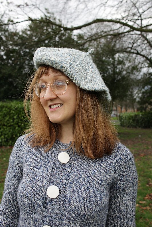 Charlotte is stood in the park wearing a blue knitted beret, and blue knitted cardigan with large round buttons
